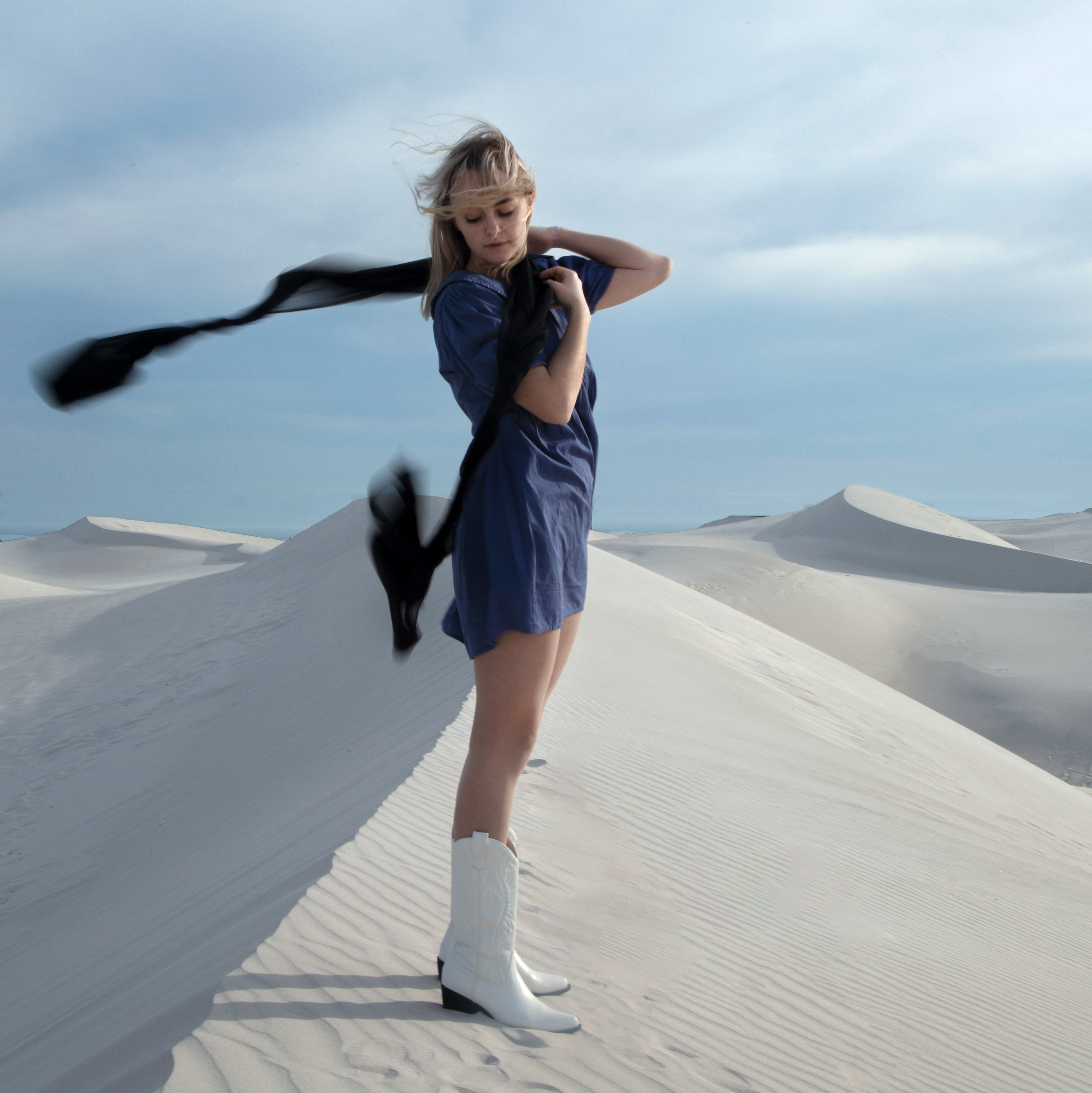 Full body photography of woman in the sand dunes with a scarf blowing in the wind and cowboy boots
